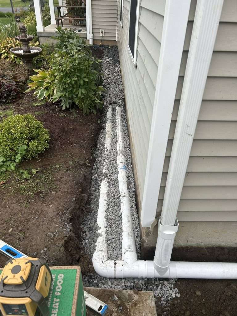 Perforated french drain pipe and a separate solid downspout line running side by side in washed stone along a house foundation in Valatie NY